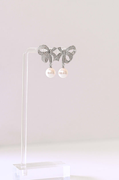 A pair of silver bow-shaped stud bridal earrings with dangling white pearls.
